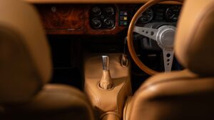 Close-up view of a classic car interior, showing tan leather seats, a wooden gear shift, a wooden steering wheel, and a dashboard with analog gauges and vintage-style controls.