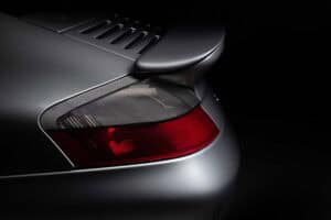 Close-up of the rear left side of a silver sports car, showing the taillight, part of the rear spoiler, and smooth metallic body lines against a dark background.