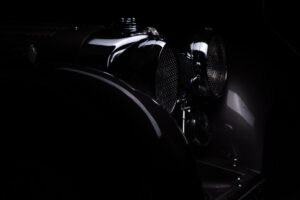 Close-up of the front of a vintage car, with curved fenders and round headlights, partially illuminated against a dark background.