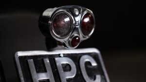 Close-up of a vintage car tail light with a shiny chrome finish and round red lenses, mounted above a black and white license plate displaying the letters "HPC" on a dark background.