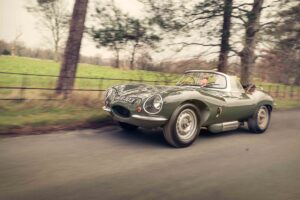 A classic green Jaguar sports car speeds down a tree-lined country road with a blurred background, emphasizing motion. The driver is visible behind the wheel, enjoying the ride.