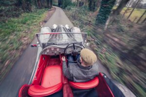 An overhead view of a person driving a vintage convertible car with red seats along a narrow, tree-lined country road. The driver wears a cap and a jacket. The background appears blurred, showing motion.