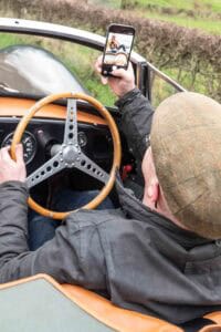 A person wearing a flat cap and glasses takes a selfie with a smartphone while sitting in the driver’s seat of a vintage convertible car with a wooden steering wheel.