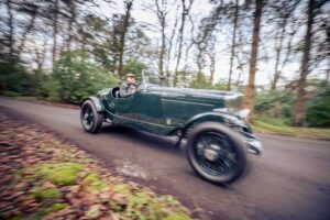 A person drives a vintage dark green Bentley convertible car along a tree-lined, leaf-strewn road. The background appears blurred, giving a sense of motion. The driver wears a cap and coat.