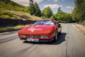 A red Ferrari sports car with the license plate X712 BKM drives on a winding country road, surrounded by green trees and hills under a partly cloudy blue sky.