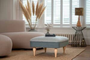 A cozy living room features a light gray ottoman with wooden legs, a tray with flowers, a beige sofa, pampas grass in a vase, a lamp, and large shuttered windows letting in natural light.