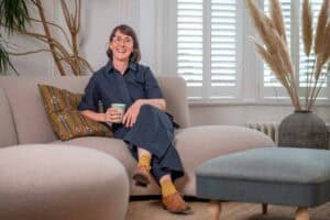A woman with glasses sits on a beige sofa, holding a mug and smiling. She is wearing a navy dress, yellow socks, and brown shoes. There are plants, a cushion, and a large window with shutters in the background.
