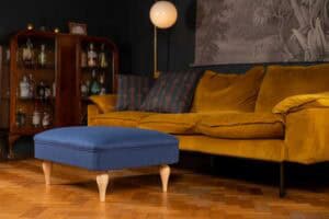 A cozy living room with a mustard yellow velvet sofa, patterned pillows, a blue ottoman with wooden legs, a vintage cabinet with bottles, parquet flooring, and a round floor lamp.