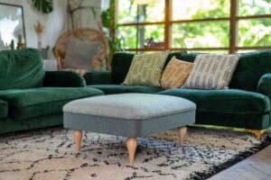 A cozy living room with a green velvet sofa, patterned cushions, a light gray ottoman, a black and white rug, and large windows showing greenery outside.