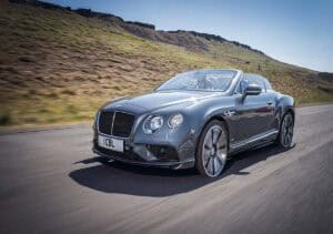 A grey Bentley convertible drives on a scenic road with grassy hills and rocky terrain in the background. The driver is visible, and the sky is clear.