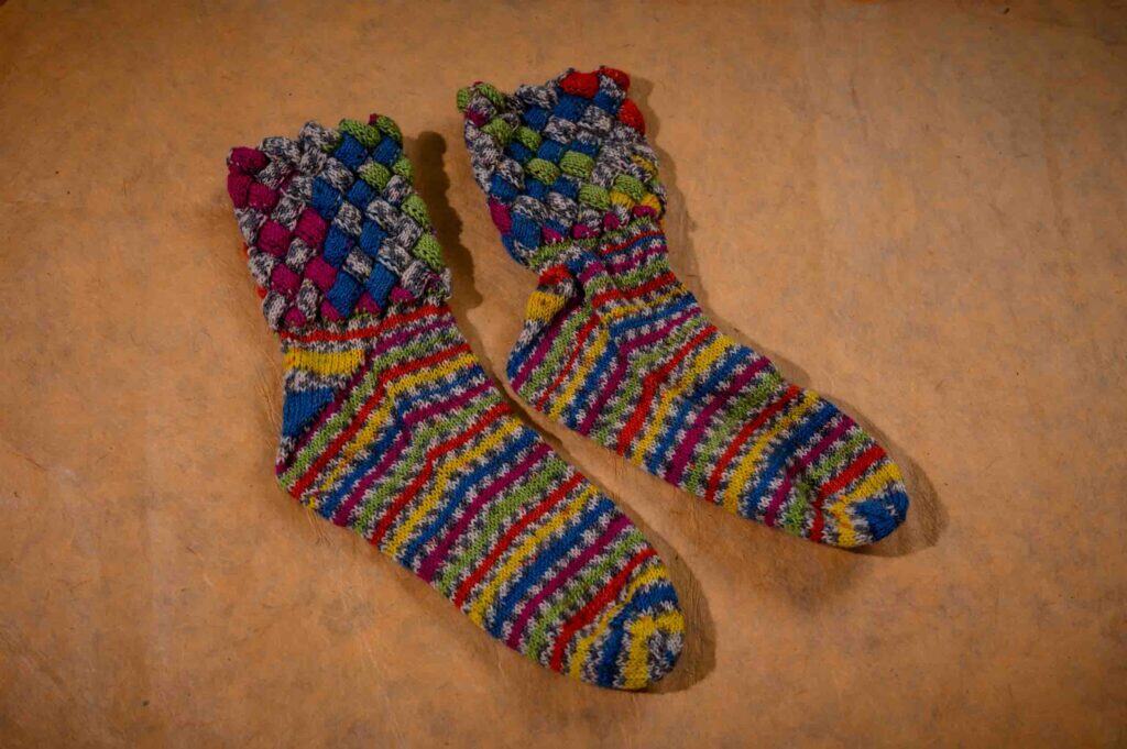 A pair of colourful, hand-knitted socks with a diamond-patterned cuff and striped patterns in shades of red, yellow, blue, green, and purple, displayed on a beige surface.