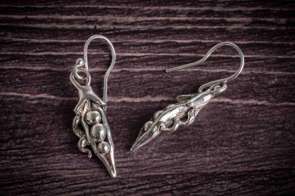 A pair of silver earrings shaped like pea pods, each with three small round beads inside, displayed on a dark wooden surface.