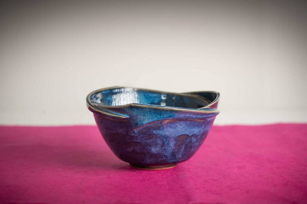 A handcrafted, deep blue ceramic bowl with a unique, asymmetrical rim sits on a pink surface against a neutral background.