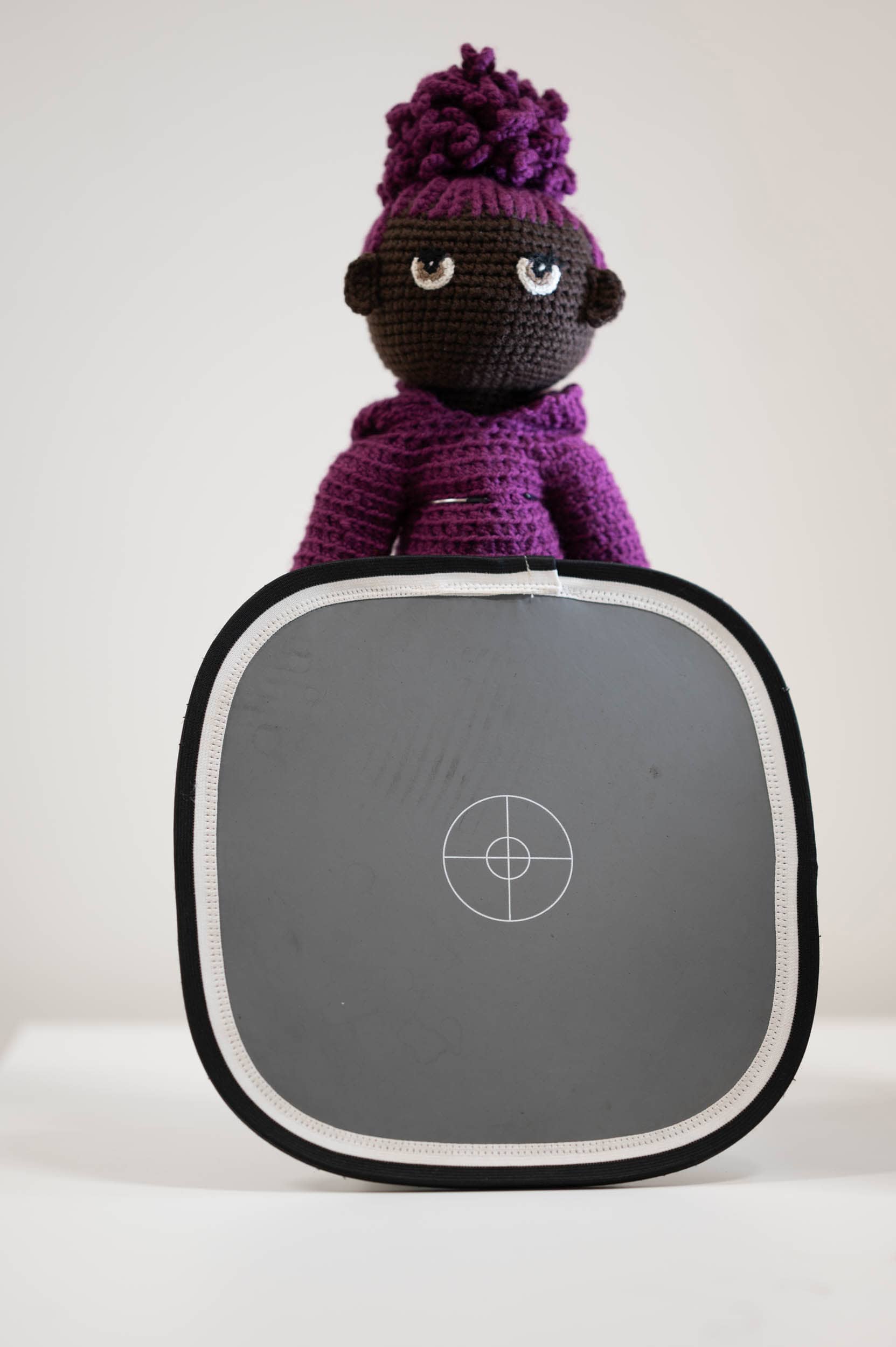 A crocheted doll with dark brown skin, a purple outfit, and big eyes sits behind an 18% grey card with a white circular target pattern, used for photography calibration, against a plain background.