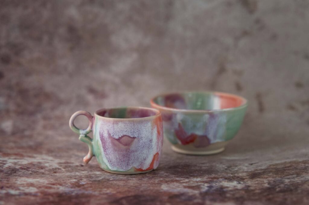 A handmade ceramic cup and bowl with a glossy, multicoloured glaze in shades of purple, green, and pink, set on a textured brown surface with a matching background.