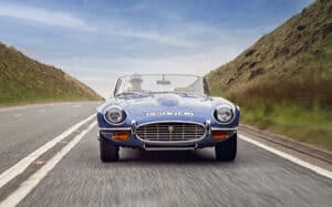 A blue classic convertible car drives down an open road between grassy hills under a blue sky. The driver wears sunglasses and a hat. The photo is taken from the front at road level.