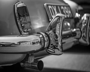 Close-up, black-and-white photo of the rear end of a vintage mrecedes car, showing shiny chrome bumper, tail light, exhaust pipe, and a partial view of a classic number plate reading "DYL 39H.