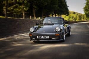 A classic black Porsche 911 turbo sports car with the number plate "DND 901Y" is being driven on a scenic, tree-lined road on a sunny day. The driver is visible behind the wheel, and the car is in motion.