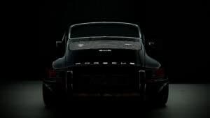 Rear view of a classic black Porsche 911S restomod by Autofarm in low light, highlighting the car’s sleek curves, chrome trim, and “Porsche” badge on the back, with much of the car fading into darkness.