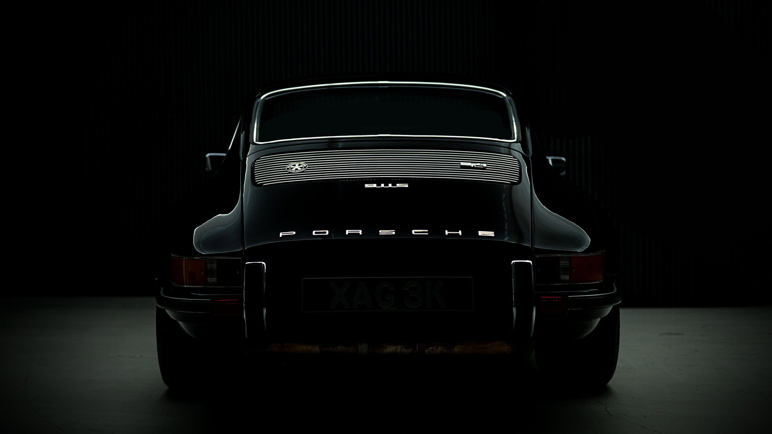 Rear view of a classic black Porsche 911S restomod by Autofarm in low light, highlighting the car’s sleek curves, chrome trim, and “Porsche” badge on the back, with much of the car fading into darkness.