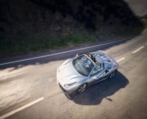 A silver convertible ferrari sports car drives along a winding road with a person behind the wheel, surrounded by blurred greenery and tarmac, suggesting speed and motion. shot from an above angle