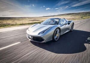 A silver ferrari sports car drives quickly along an empty, scenic road with grassy fields and blue sky in the background.