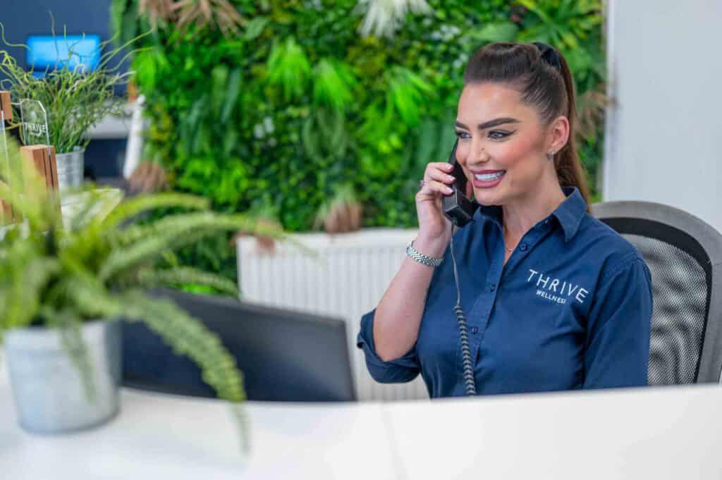 A woman in a "Thrive Wellness" shirt smiles whilst speaking on the phone at a modern reception desk, with green plants and a computer screen visible in the background.