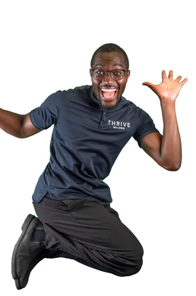 A man wearing glasses, a navy "THRIVE" polo shirt, and black trousers jumps excitedly with a wide smile and raised hands against a white background.