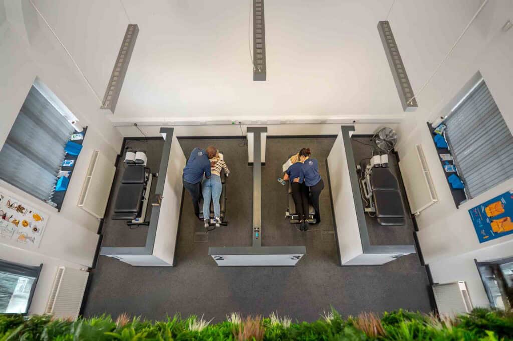 Overhead view of two patients lying on separate treatment couches in a clinic, each attended by a practitioner. The room is divided by partitions, with medical posters and equipment visible.