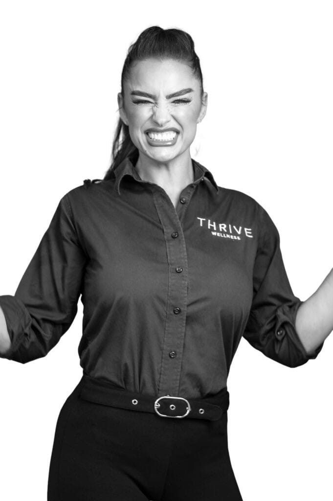 Black and white photo of a woman smiling widely with her eyes closed, wearing a button-up shirt that reads "THRIVE WELLNESS." Her hair is up in a ponytail and her arms are raised in excitement.