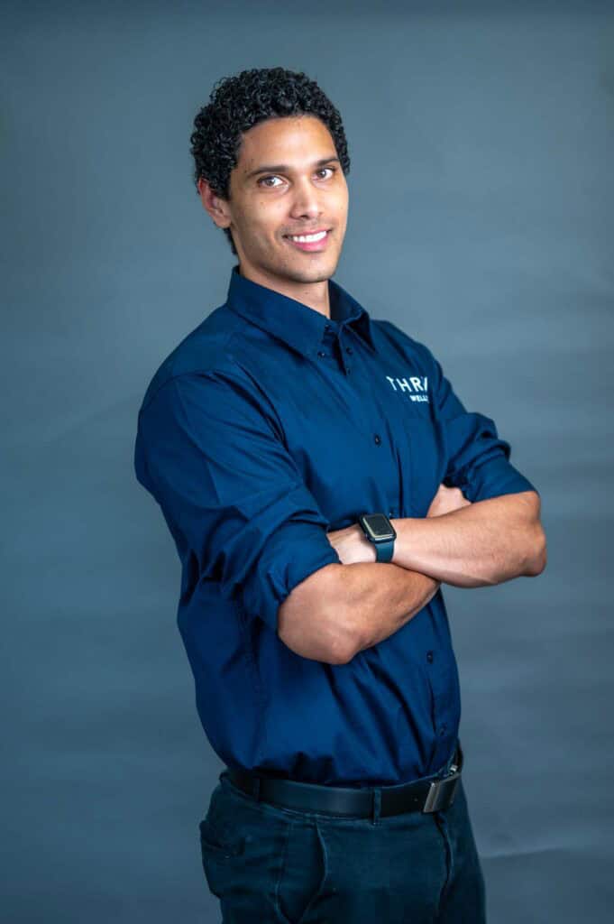 A man with curly hair stands with arms crossed, wearing a navy blue shirt and black trousers, smiling slightly against a plain grey background. He is also wearing a smartwatch.