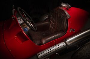 Close-up view of the cockpit of a vintage red ERA racing car, showing a worn brown leather seat, a steering wheel, and part of the car’s body and exhaust pipe, with dark surroundings.