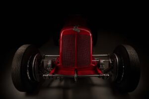 A dramatic, front-facing image of a vintage red racing ERA car with large exposed wheels, chrome suspension, and a mesh grille, emerging from a dark background. Shot using the FDL photographic technique