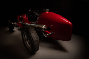 A vintage red racing ERA car photographed in low light, highlighting its curved rear, large exposed exhaust pipe, and textured rear tyre on a smooth grey floor. Shot using the FDL photographic technique