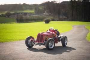 A vintage red ERAracing car with exposed wheels sits on a winding tarmac road, surrounded by bright green grass and trees, with buildings and hills in the background under a clear sky.