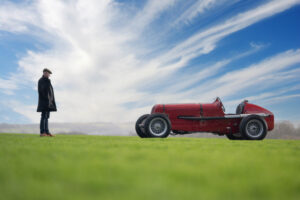 Tom Hardman in a dark coat and hat stands on grass, facing a vintage red ERA racing car under a blue sky with wispy clouds.