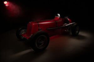 A vintage red ERA racing car is dramatically lit from the side and rear, highlighting its sleek curves and large exposed wheels, with dark shadows and a red spotlight in the background. Shot using the FDL photographic technique