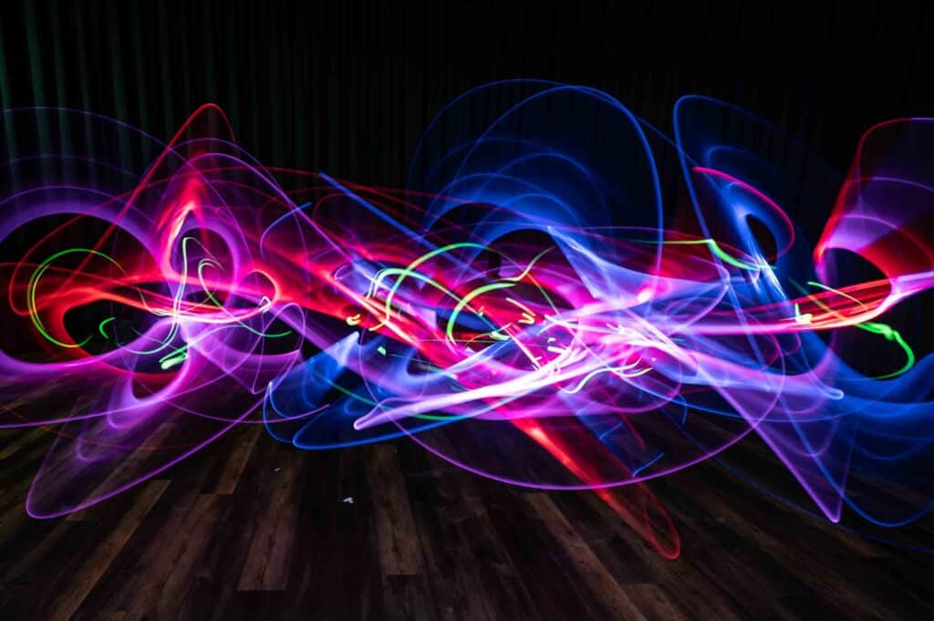 Swirling trails of vibrant red, blue, purple, and green light form abstract patterns over a dark background and wooden floor, creating a dynamic, energetic effect.