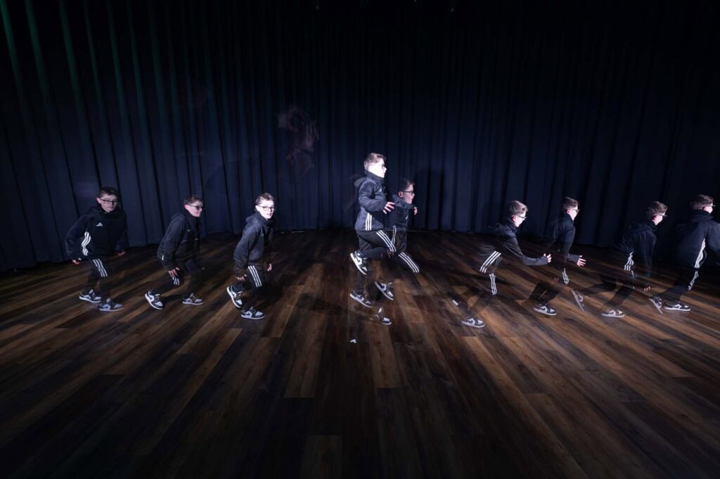 A sequence of overlapping images shows a boy in a black tracksuit jumping in stages across a wooden floor, against a dark curtain background, illustrating movement.