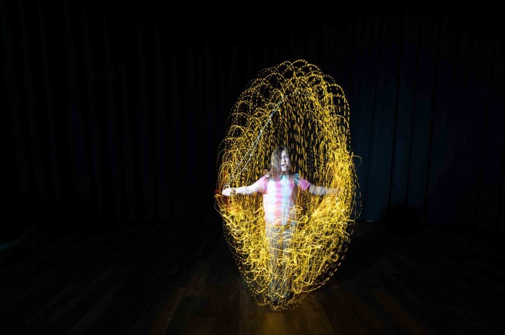 A child stands in a dark room, surrounded by swirling yellow light trails, creating an illuminated, dome-like shape around them. The child is smiling and wearing a striped jumper.