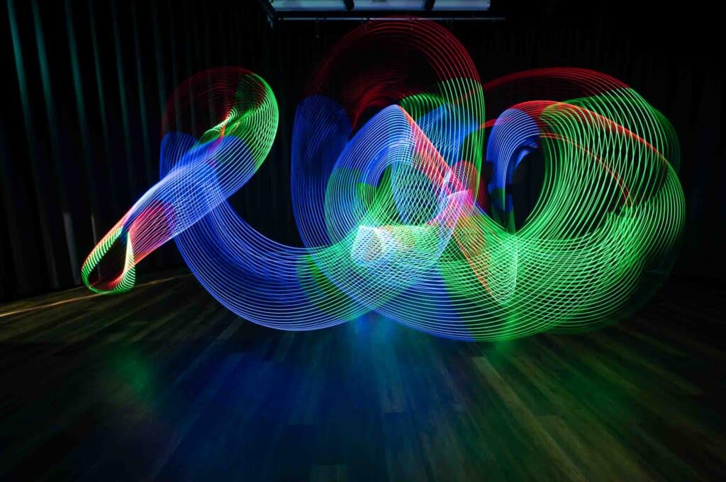 Colourful light trails of blue, green, and red swirl and overlap in dynamic patterns against a dark room with wooden flooring and black curtains.