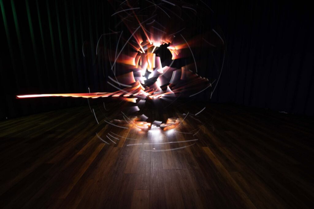 A long-exposure photo shows swirling, bright light trails forming a dynamic, abstract pattern above a wooden floor in a dark room with black curtains.