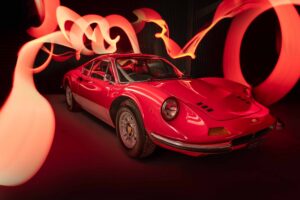 A classic red sports car is parked indoors with sweeping trails of red and orange light swirling artistically around it, creating a dynamic and dramatic atmosphere.