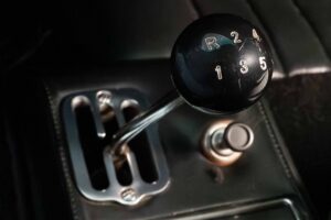 Close-up of a manual ferrari dino car gear lever with a black knob showing 5 speeds and reverse, next to a metallic gating pattern and a small round button on a dark car interior. shows the H pattern of a ferrari gearbox