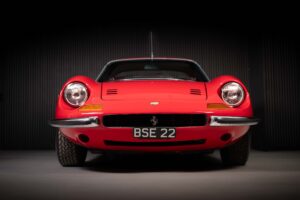 A front view of a classic red Ferrari dino sports car with round headlamps, a chrome bumper, and a number plate reading "BSE 22," set against a dark, minimalist background.