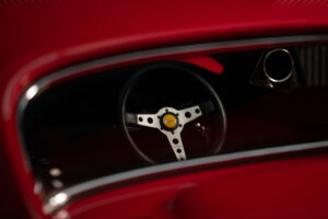 Close-up view through a car window showing a classic black and silver steering wheel with a yellow centre emblem, surrounded by a vibrant red car interior.