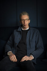 A man with short curly hair and glasses sits on a dark sofa, wearing a navy jacket, black jumper, and white shirt. He looks calmly at the camera, with his hands resting in his lap against a dark background.