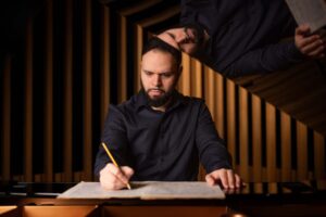A bearded man in a dark shirt writes on sheet music with a pencil at a piano. His reflection is visible on the piano's glossy surface behind him. Vertical wooden panels form the background.