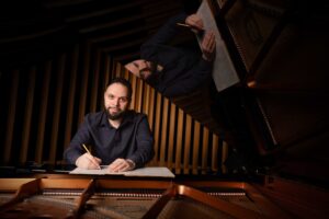 A man sits at a piano, writing music on manuscript paper with a pencil. His reflection is visible on the raised piano lid above him. The background features vertical wooden panels.