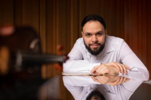 A man with a beard, wearing a white shirt, leans on a shiny surface reflecting his face. He looks at the camera and a violin is blurred in the foreground. The background features vertical wooden panels.